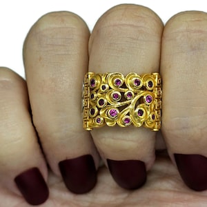 May include: A gold ring with a delicate, intricate design featuring swirls and small pink gemstones. The ring is worn on a finger with dark red nail polish.