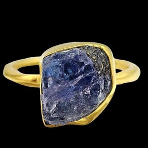 Puede incluir: Un anillo de oro con una gran piedra preciosa de tanzanita en bruto de color azul intenso, engastada en un bisel liso de forma orgánica. La piedra muestra una textura cristalina rugosa con una inclusión de tono metálico oscuro.