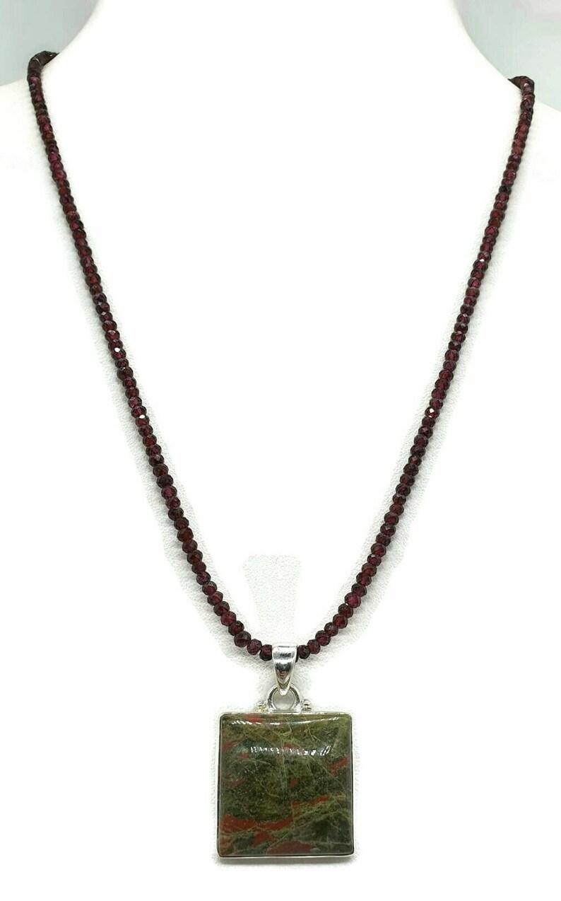 May include: A necklace made of small, dark red beads with a square pendant featuring a green and red stone.