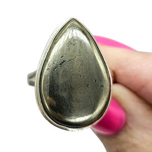 May include: A ring featuring a large, teardrop-shaped pyrite gemstone with a metallic, brassy gold luster and natural surface texture, securely set within a smooth, polished silver-toned metal bezel that frames the stone's unique shape.