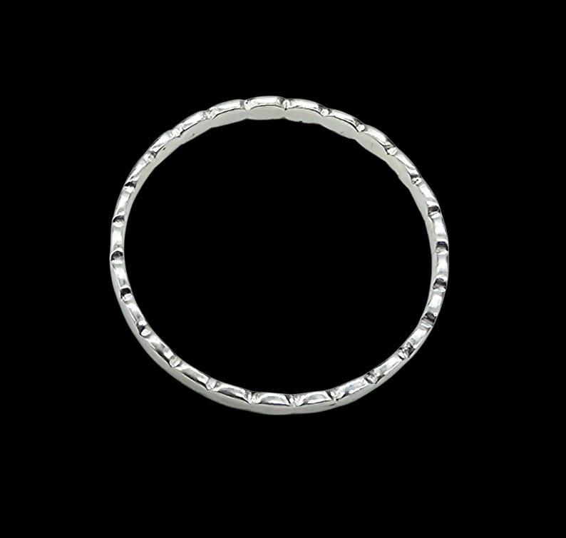 Puede incluir: Un anillo plateado con un patr&oacute;n repetitivo de peque&ntilde;as hendiduras curvas alrededor de la banda. El anillo est&aacute; sobre un fondo negro, resaltando su brillo met&aacute;lico y su intrincado dise&ntilde;o. El dise&ntilde;o del anillo es simple pero elegante.