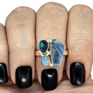 May include: A silver ring featuring three opal stones. The stones are a dark blue, a light blue, and a green opal. The ring is worn on a finger with black nail polish.