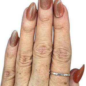 Puede incluir: Primer plano de una mano con u&ntilde;as largas en forma de almendra pintadas de un color marr&oacute;n claro brillante. Un anillo plateado con un dise&ntilde;o ovalado repetido est&aacute; en el dedo anular. Las u&ntilde;as tienen un acabado brillante.