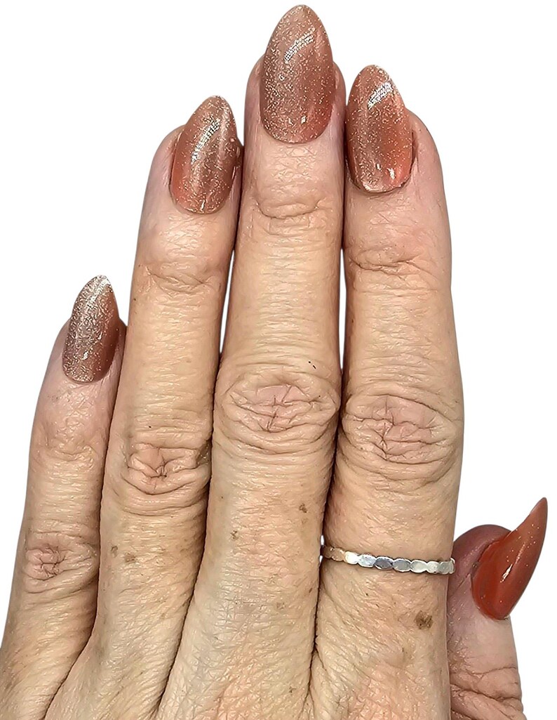 Puede incluir: Primer plano de una mano con u&ntilde;as largas en forma de almendra pintadas de un color marr&oacute;n claro brillante. Un anillo plateado con un dise&ntilde;o ovalado repetido est&aacute; en el dedo anular. Las u&ntilde;as tienen un acabado brillante.