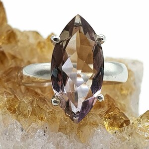 May include: A marquise-shaped, light brown gemstone ring set in sterling silver. The ring is displayed on a bed of small, golden crystals.