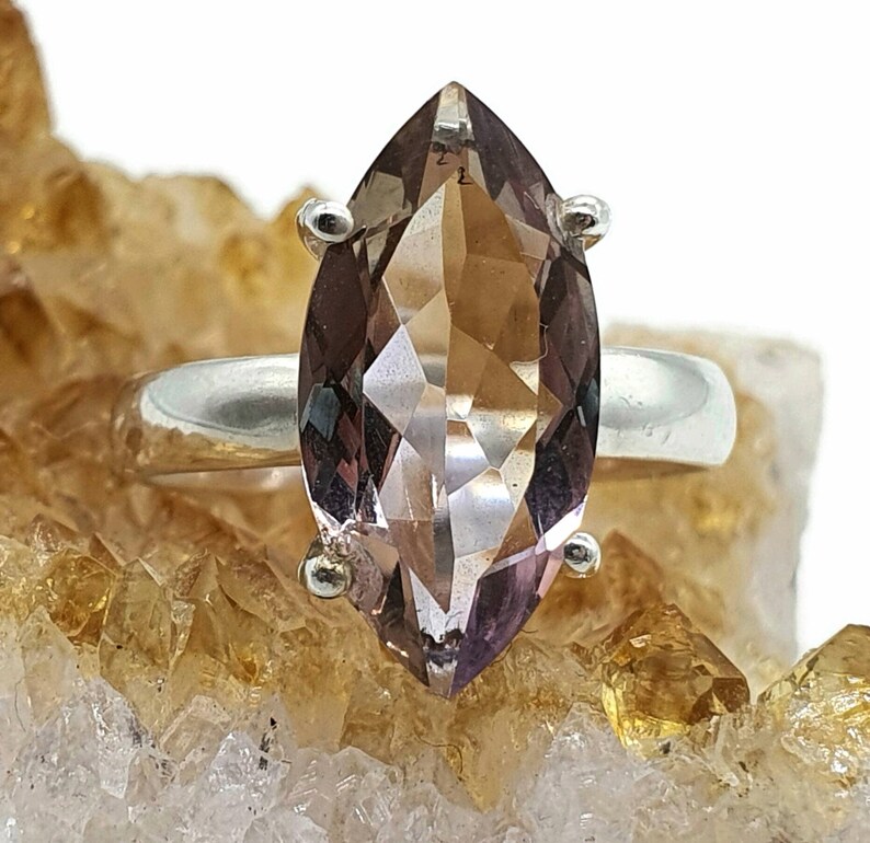 May include: A marquise-shaped, light brown gemstone ring set in sterling silver. The ring is displayed on a bed of small, golden crystals.