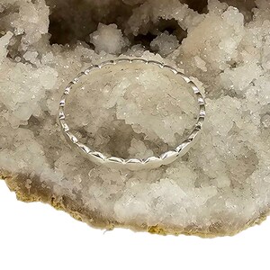 Puede incluir: Un anillo de plata con borde festoneado, sobre una geoda de cristal blanca y beige. El dise&ntilde;o del anillo presenta una serie de formas redondeadas y repetitivas, creando una apariencia delicada y texturizada. El anillo es una joya.