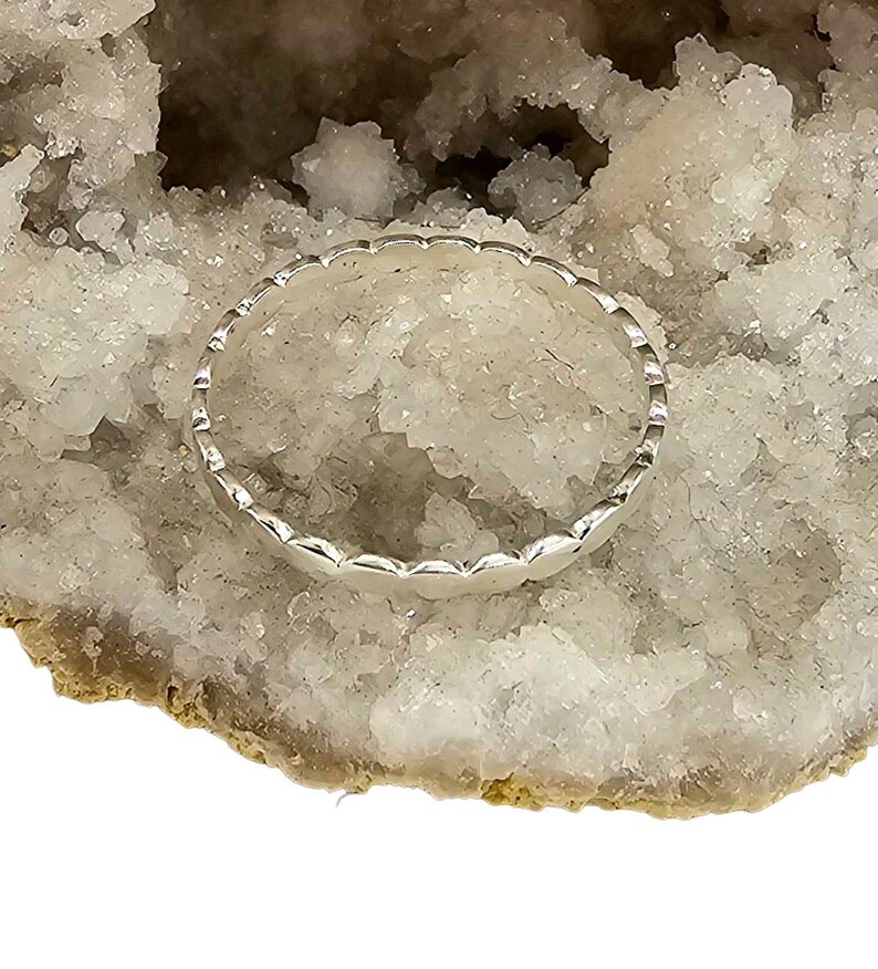 Puede incluir: Un anillo de plata con borde festoneado, sobre una geoda de cristal blanca y beige. El dise&ntilde;o del anillo presenta una serie de formas redondeadas y repetitivas, creando una apariencia delicada y texturizada. El anillo es una joya.