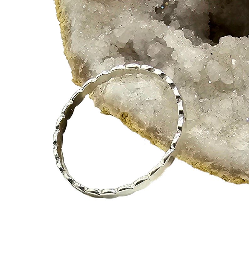 Puede incluir: Un anillo de plata con un dise&ntilde;o de borde festoneado. El anillo es circular y parece estar hecho de un metal brillante. El anillo se muestra sobre un fondo blanco y beige.
