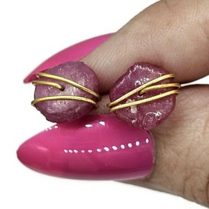 May include: A pair of handcrafted earrings featuring rough, pink gemstones wrapped in gold-colored wire. The earrings have a natural, organic look, showcasing the unique texture and color of the stones.