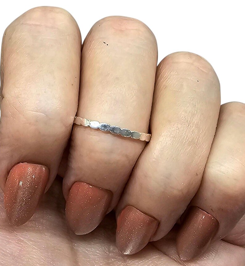 Puede incluir: Un anillo plateado con un dise&ntilde;o festoneado repetido. El anillo se lleva en un dedo con u&ntilde;as pulidas de color marr&oacute;n rojizo. El delicado dise&ntilde;o del anillo a&ntilde;ade un toque de elegancia a la mano.