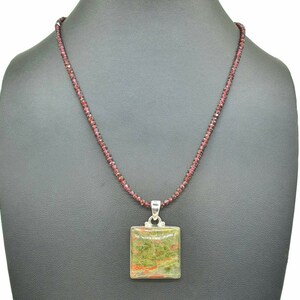 May include: A silver necklace with a square green, red, and brown stone pendant. The necklace is made of small red beads.