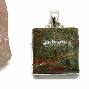 May include: A square pendant with a green and red jasper stone set in sterling silver. The stone has a mottled pattern of green, red, and brown.