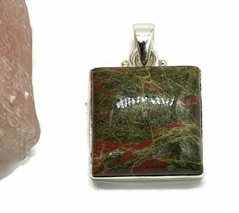 May include: A square pendant with a green and red jasper stone set in sterling silver. The stone has a mottled pattern of green, red, and brown.