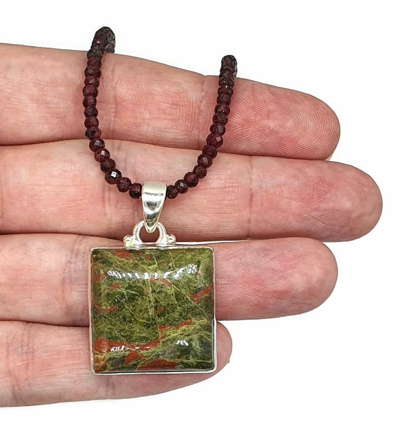 May include: A silver pendant necklace with a square green and red jasper stone. The necklace is made of small, dark red beads.