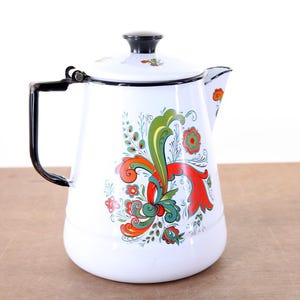 May include: White enamelware coffee pot with a black handle, lid, and trim. The pot is decorated with a colorful floral design in red, green, and orange. A vintage kitchenware item.