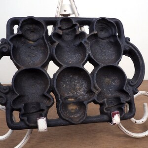 May include: Cast iron mold for making snowman-shaped treats. The mold has six individual cavities, each shaped like a snowman with a hat. The mold is black and has a rustic appearance.