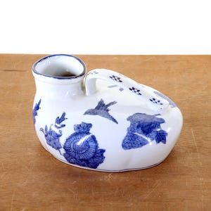 May include: A white ceramic chamber pot with blue floral and bird designs. The pot has a handle and a spout. The design includes a bird in flight and floral patterns. The pot is on a wooden surface.