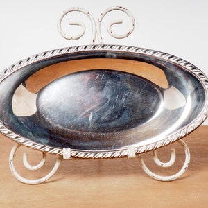 May include: A silver oval serving dish with a rope-like design around the edge, sitting on a white metal stand with decorative scrolls.