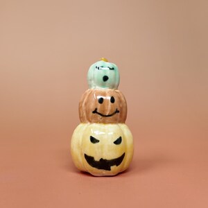 May include: A ceramic stack of three pumpkins in a vertical arrangement. The top pumpkin is light green with a surprised face, the middle is brown with a smiling face, and the bottom is yellow with a jagged, grinning face, all painted in black.