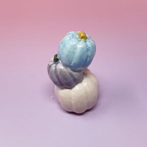 Peut inclure: Une pile de trois citrouilles en céramique avec une finition brillante. La citrouille du haut est bleu clair avec une tige jaune, celle du milieu est violette avec une tige verte, et celle du bas est d'une couleur blanc rosé pâle.