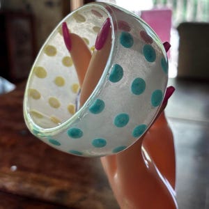 May include: A wide, translucent bangle bracelet featuring a pattern of embedded polka dots in alternating shades of light blue and pale yellow. The bracelet has a smooth, glossy finish and a rounded, thick profile that catches the light.