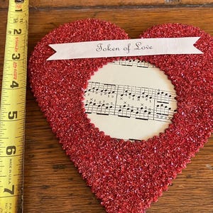 May include: A red glitter-covered heart-shaped decoration with a central opening revealing sheet music. A banner across the top reads "Token of Love." The heart is approximately 6 inches wide. A Stanley measuring tape is in the image.