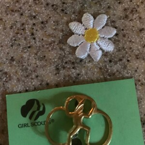 May include: A gold-colored Girl Scout Brownie pin with the Girl Scouts logo and the text "Girl Scouts" on a green card with the text "Brownie Girl Scout Pin".