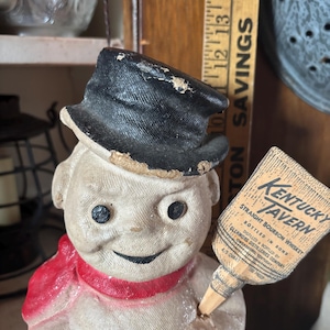 May include: A vintage snowman figurine with a black top hat and a red scarf. The snowman holds a paper advertisement for Kentucky Tavern Straight Bourbon Whiskey. The figurine has black button eyes and a smiling face.