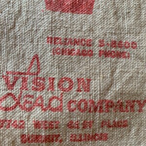 May include: Close-up of a beige burlap sack with red printed text. The text includes "VISION BEAD COMPANY" and an address in Illinois. The burlap has a rough, textured surface.