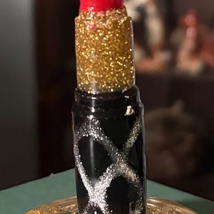 May include: A decorative lipstick-shaped object with a vibrant red tip, a gold glitter midsection, and a black base adorned with silver glitter designs. It rests on a gold-flecked, clear resin base, creating a glamorous aesthetic.