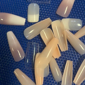 May include: A collection of long, oval-shaped, false fingernails in various shades of pink, white, and clear. The nails are arranged on a blue surface.