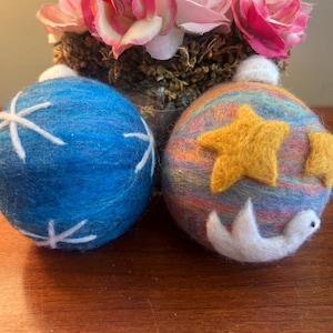 Op de afbeelding: Twee handgemaakte vilten kerstballen. Eén is blauw met witte sterrenpatronen. De andere is veelkleurig met gele sterren en een wit vogelontwerp. Roze rozen staan op de achtergrond.
