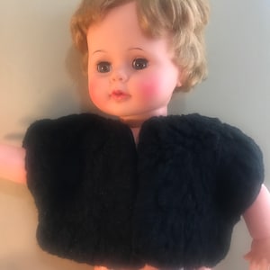 Könnte beinhalten: Eine Vintage-Puppe trägt eine schwarze Kunstfelljacke. Die Puppe hat blondes Haar, blaue Augen und rosige Wangen.