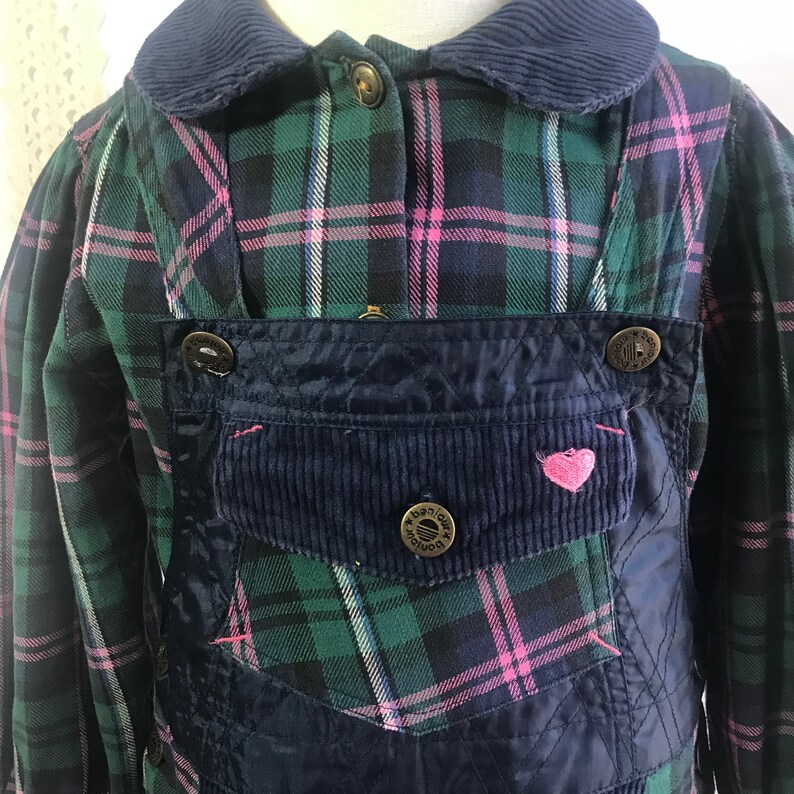 May include: A blue and green plaid shirt with a corduroy collar and a blue overall dress with a heart-shaped patch. The overall dress has a quilted design and a pocket with a button that says "Banjo"
