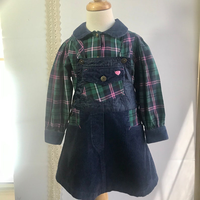 May include: A green and pink plaid shirt dress with a navy blue corduroy overlay. The dress has a Peter Pan collar and a small pink heart on the front.
