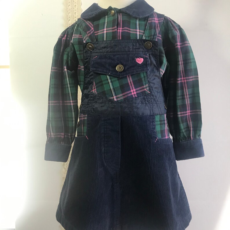 May include: A blue corduroy overall dress with a green and pink plaid shirt. The dress has a large pocket on the front with a small pink heart.