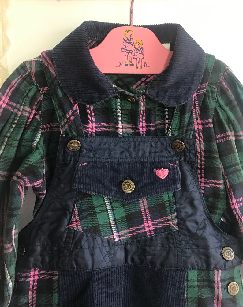 May include: A blue overall dress with a green and pink plaid shirt. The overall dress has a corduroy patch pocket with a pink heart on it. The shirt has a navy blue corduroy collar.