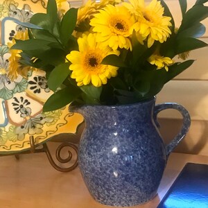 May include: A blue ceramic pitcher with a speckled glaze, filled with a bouquet of yellow daisies.
