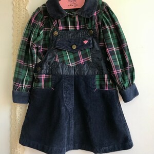 May include: A green and pink plaid shirt with a navy blue corduroy overall dress. The dress has a heart-shaped patch on the front and two pockets on the sides.