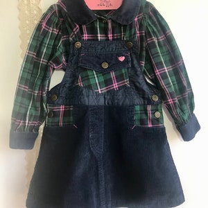 May include: A green and pink plaid shirt with a navy blue corduroy overall dress. The dress has a heart-shaped patch on the front and a small pocket on the chest.