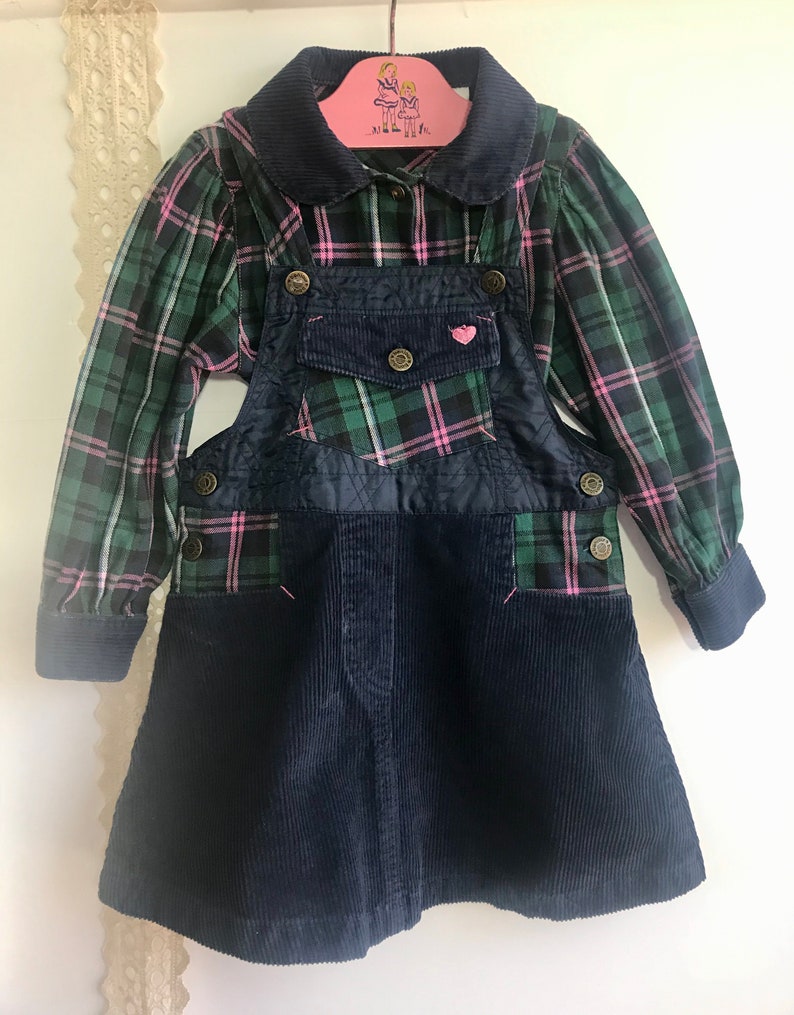 May include: A green and pink plaid shirt with a navy blue corduroy overall dress. The dress has a heart-shaped patch on the front and a small pocket on the chest.