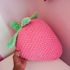 May include: A pink crocheted strawberry pillow with green leaves. The pillow is soft and plush, perfect for cuddling.