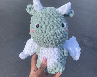 Puede incluir: Un dragón de peluche tejido a crochet con pelaje verde y blanco. El dragón tiene ojos grandes, una pequeña nariz rosa y alas blancas.