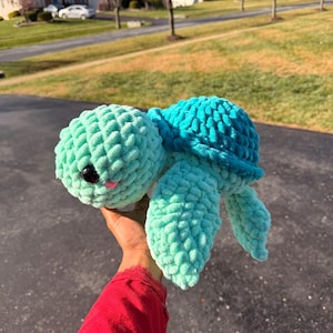 May include: A handmade plush turtle in shades of teal and turquoise. The turtle has a textured, chunky knit appearance, with a round head, flipper-like limbs, and a shell. It features black eyes and a pink mouth. A whimsical, soft toy.