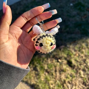 May include: A crocheted bee keyring with black and yellow chequered pattern, white wings, and pink cheeks. The bee is attached to a silver key ring.