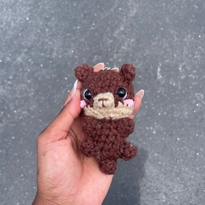 May include: A brown crocheted bear keychain with a light brown nose and pink cheeks.