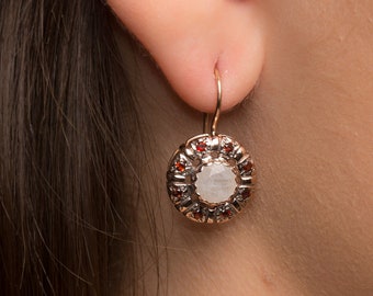 May include: A rose gold earring with a circular design. The center features a white stone, surrounded by small red stones and a rose gold frame. The earring is suspended from a gold hook.