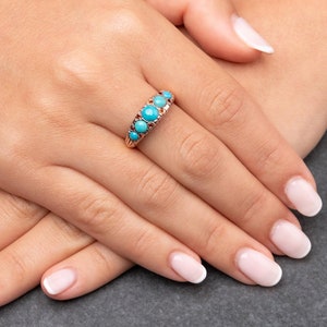 Pode incluir: Um anel de ouro com pedras turquesa engastadas em fila. O anel está no dedo de uma pessoa.