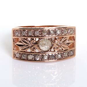 May include: A rose gold ring with a large, clear, round gemstone surrounded by smaller, clear gemstones. The ring has a decorative, ornate design.